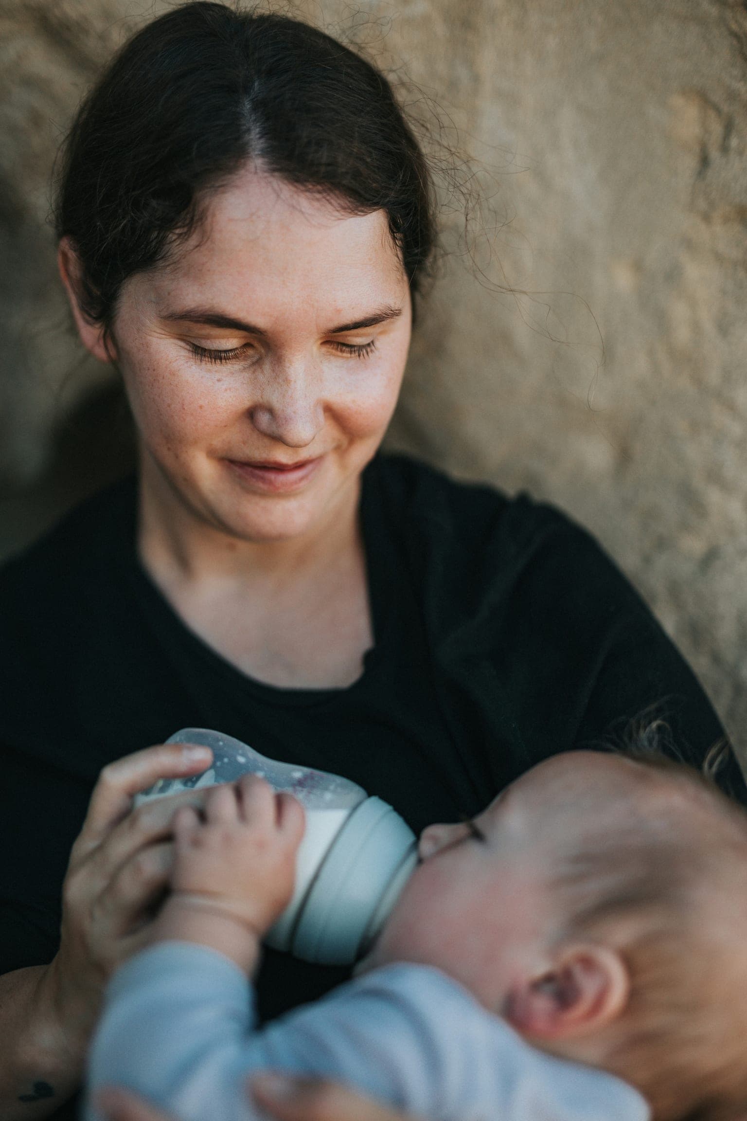 Mother smiling while bottle-feeding her baby outdoors against a soft stone wall