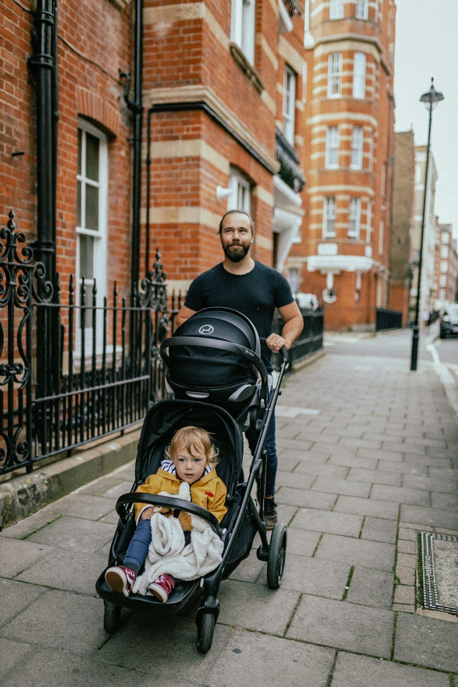 Father pushing a double stroller with a toddler in a yellow raincoat along a city sidewalk