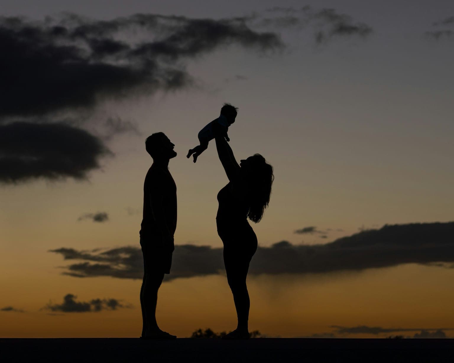 Silhouette of parents lifting their child against a warm sunset sky